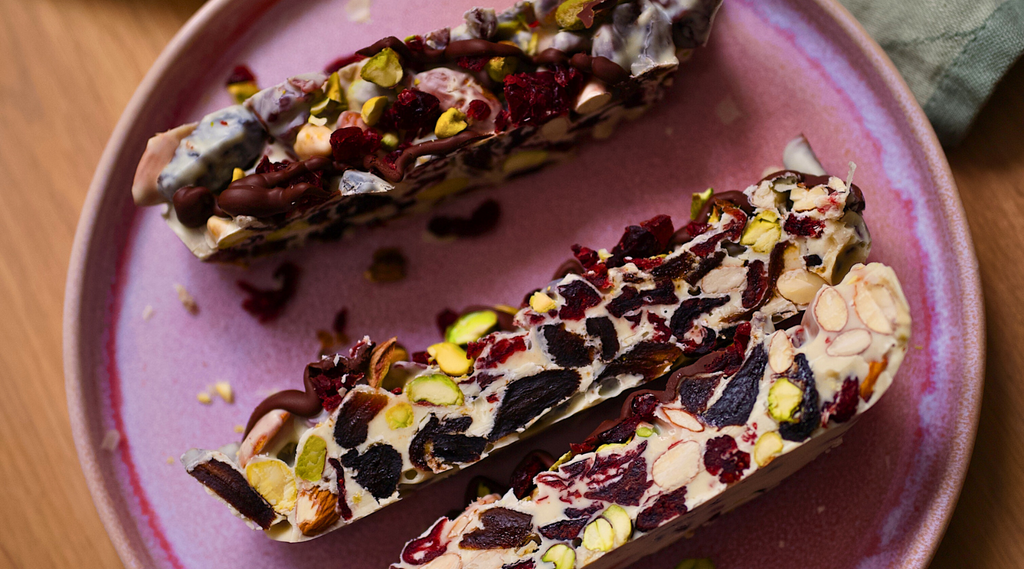 Stücke von hausgemachtem Rocky Road mit weißer Schokolade, garniert mit Mandeln, getrockneten Aprikosen, ungesalzenen Pistazien, Cranberries und Kirschen auf einem Steingutteller.